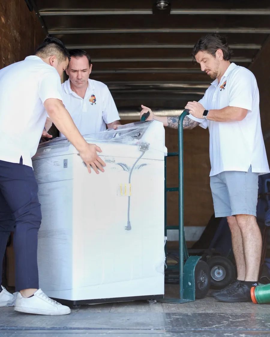 Stack Haus movers loading a wrapped washing machine onto a truck with a hand truck